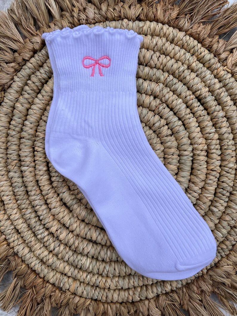 May include: White ankle socks with a pink embroidered bow on the top. The socks have a ribbed texture and a ruffled edge.