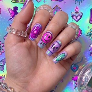 Puede incluir: Una mano con u&ntilde;as largas de forma cuadrada con dise&ntilde;os vibrantes en morado, rosa y azul que incluyen una espiral, una cara sonriente que se derrite, una mariposa y un cristal. La mano lleva tres anillos de pl&aacute;stico transparente y una pulsera.