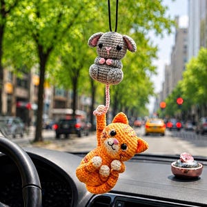Op de afbeelding: Een gehaakte auto-achteruitkijkspiegelhanger met een kleine grijze muis met roze accenten die boven een oranje cyperse kat hangt. De kat houdt met één poot de lange roze staart van de muis vast en beide figuren hebben zwarte kraaloogjes.