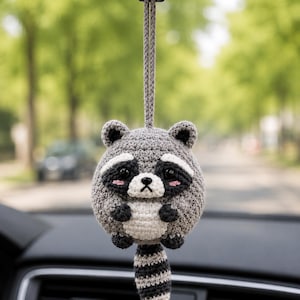 May include: A round, crocheted raccoon ornament hanging from a gray cord. The raccoon features a gray body, a white belly, black eye patches, small black ears, and a striped tail. It has pink blush marks on its cheeks and black bead eyes.