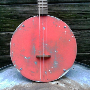 May include: A three-stringed banjo with a circular, distressed red body featuring visible paint wear and metal screws. The instrument has a wooden neck and bridge, with thin strings running vertically across the center of the round face.