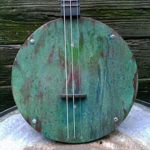 May include: A circular banjo body featuring a weathered green patina finish with streaks of rust-colored brown and blue paint splatters. The instrument has three vertical strings, a dark bridge, and four silver-toned metal screws securing the face.