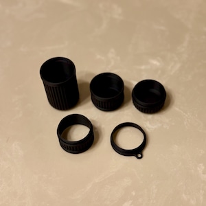 May include: A selection of black plastic bottle caps and closures. The set includes a tall cylindrical piece, three circular caps, and a ring with a small tab. The items are arranged on a light-coloured surface.