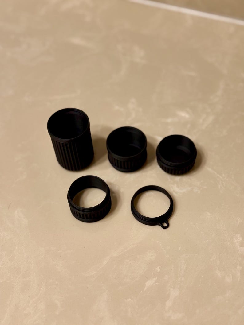 May include: A selection of black plastic bottle caps and closures. The set includes a tall cylindrical piece, three circular caps, and a ring with a small tab. The items are arranged on a light-coloured surface.