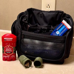 May include: A black travel bag with various compartments, a red deodorant stick, a tube of toothpaste, and two green pill containers. The deodorant is labelled "Old Spice Sweat Defense Swagger". The toothpaste is labelled "Advanced Deep Clean Mint".