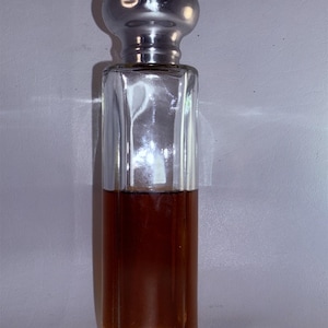 Puede incluir: Una botella de vidrio transparente con una tapa plateada y un líquido marrón en su interior. La botella tiene una forma rectangular y un fondo ligeramente redondeado.