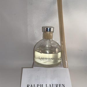 Peut inclure: Un flacon en verre transparent de parfum d'intérieur Ralph Lauren avec un bouchon argenté et des bâtonnets en bois. Le flacon contient un liquide clair et l'étiquette indique "LAUREN RALPH LAUREN". Une carte blanche avec "RALPH LAUREN HOME FRAGRANCE" est devant.