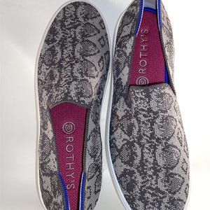 Pode incluir: Um par de sapatos cinza e branco de deslizamento com um padrão de pele de cobra. Os sapatos têm um forro rosa com a palavra "ROTHYS" impressa no interior.