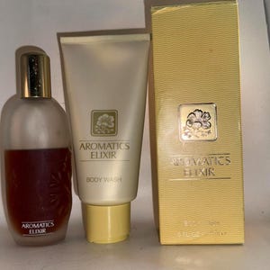 Może przedstawiać: Zestaw zapachowy Aromatics Elixir z matową szklaną butelką ze złotą zakrętką, tubką żelu pod prysznic i złotym pudełkiem. Nazwa marki i produktu są wydrukowane.