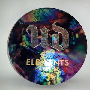 May include: Round eyeshadow palette with a colorful, marbled design. The palette features the text "ELEMENTS PALETTE" in gold lettering, with a silver logo at the top. The palette has a holographic effect.