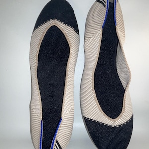 Puede incluir: Un par de bailarinas beige y negras. La puntera y el talón son negros, mientras que el resto del zapato es beige. El ribete es beige con un acento azul. Los zapatos están hechos de un material tejido.