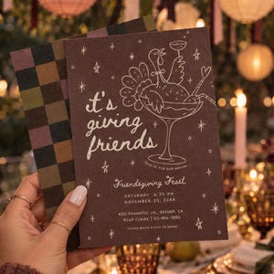 Whimsical Friendsgiving dinner invitation held in front of a warmly lit outdoor Thanksgiving tablescape with glowing candles and autumn decorations. The deep brown invitation features blush pink illustrated cocktail glass artwork, delicate stars, bows, and playful “it's giving friends” lettering. This customizable Canva fall party invitation is designed for a friends Thanksgiving dinner, girls night potluck, or autumn gathering and can be printed or sent as a digital invite.