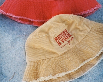 May include: Two corduroy bucket hats with frayed edges. The top hat is red with white text that reads "Rhyder & Vixen - swim". The bottom hat is beige with red text that reads "Rhyder & Vixen - swim".