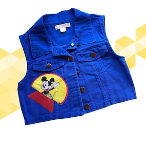May include: A blue denim vest with a Mickey Mouse graphic on the front. The vest has a collared neckline, button closures, and two front pockets. The Mickey Mouse graphic features Mickey in a yellow circle and red triangle.
