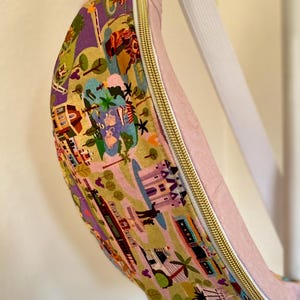 May include: A pink fanny pack with a gold zipper and a white strap. The main body of the bag features a colorful print with illustrations of buildings, trees, and people. The bag is curved and appears to be made of fabric.