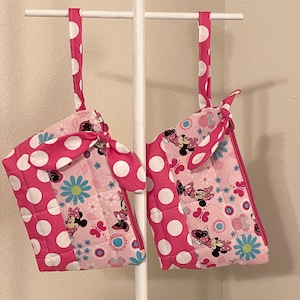 May include: Two pink oven mitts with white polka dots and Minnie Mouse designs hang from a white rack. The mitts have a quilted pattern and feature blue flowers and butterflies.
