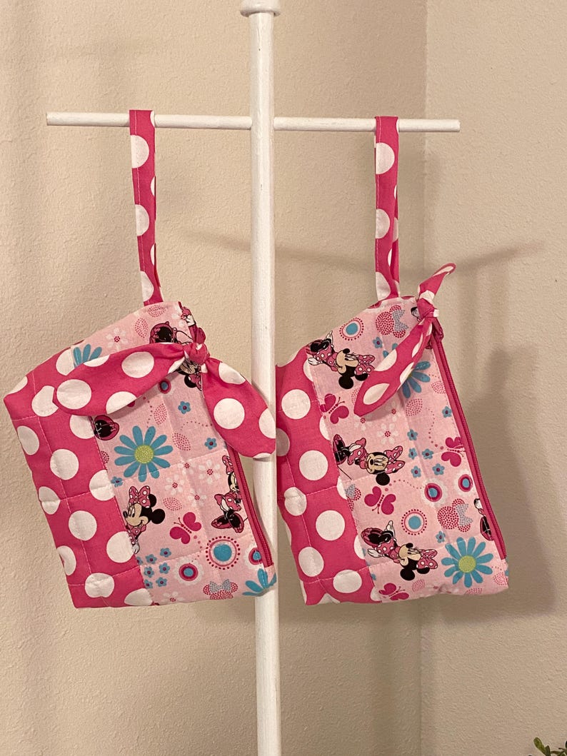 May include: Two pink oven mitts with white polka dots and Minnie Mouse designs hang from a white rack. The mitts have a quilted pattern and feature blue flowers and butterflies.