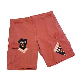 May include: Red cargo shorts with Mickey Mouse patches on the pockets. The shorts have a button closure and multiple pockets. The patches feature Mickey Mouse and star designs with red and white stripes.