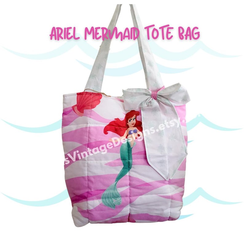 May include: A pink and white quilted tote bag featuring a cartoon image of Ariel the mermaid. The bag has white handles and a large bow. Text at the top reads "ARIEL MERMAID TOTE BAG".