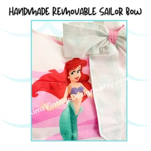May include: Handmade removable sailor bow featuring a cartoon mermaid with red hair, a purple top, and a green tail. The bow is white with a light blue pattern and a pink center. The text "Handmade Removable Sailor Bow" is at the top.
