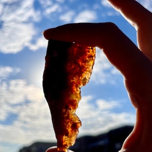 May include: A translucent, amber-colored stone with a rough, textured surface is held against a bright blue sky with scattered clouds. The stone's edges are irregular, and the sunlight shines through it, creating a warm glow.