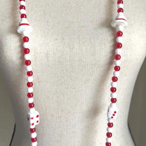 Peut inclure: Long collier composé de perles alternées rouges et blanches. Le collier comporte des breloques blanches en forme de dés avec des points rouges et des breloques blanches en forme de champignon. Un pendentif en forme de cœur doré avec un motif rouge et blanc est suspendu en bas.