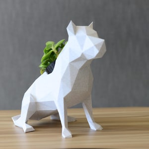 May include: A white geometric dog planter with a small green plant in its back. The dog is made of folded paper or cardboard, with a faceted design. The planter sits on a light brown wooden surface against a gray background.