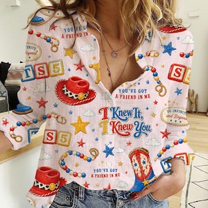 May include: A long-sleeved button-down shirt in white featuring a colorful repeating pattern of red cowboy hats, blue cowboy boots, yellow stars, clouds, beaded strings, and blocks with the text TS5. The text reads You've Got A Friend In Me.