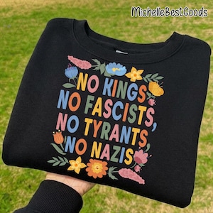 Peut inclure: Sweat-shirt noir avec l'inscription multicolore "NO KINGS, NO FASCISTS, NO TYRANTS, NO NAZIS", entourée de motifs floraux. Le sweat-shirt est tenu par une personne.