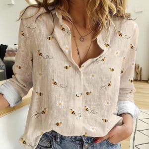 May include: Beige button-down shirt with a bee and daisy pattern. The long-sleeved shirt has a collared neckline and is made of a lightweight fabric. The cuffs are a light blue color. The shirt is unbuttoned at the top.