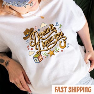 K&ouml;nnte beinhalten: Ein wei&szlig;es T-Shirt mit einem grafischen Design und dem Text "I knew it, I knew you" in einer seilartigen Schriftart. Das Design enth&auml;lt einen braunen Cowboyhut, ein gelbes Hufeisen, einen bunten Buchstabenw&uuml;rfel mit den Buchstaben Y und D sowie Sterne.