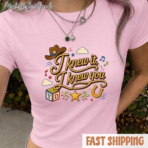 K&ouml;nnte beinhalten: Ein hellrosa T-Shirt mit kurzen &Auml;rmeln und einem Grafikdruck mit dem Text "I knew it, I knew you" in einer seilartigen Schriftart. Das Design umfasst einen braunen Cowboyhut, einen bunten Buchstabenw&uuml;rfel, einen gelben Stern und ein Hufeisen.