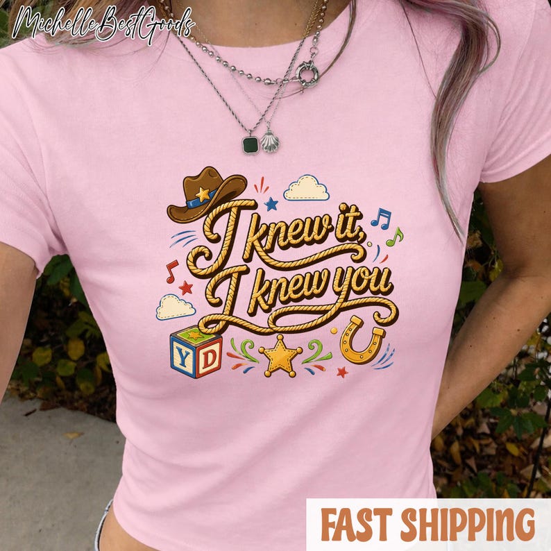 K&ouml;nnte beinhalten: Ein hellrosa T-Shirt mit kurzen &Auml;rmeln und einem Grafikdruck mit dem Text "I knew it, I knew you" in einer seilartigen Schriftart. Das Design umfasst einen braunen Cowboyhut, einen bunten Buchstabenw&uuml;rfel, einen gelben Stern und ein Hufeisen.