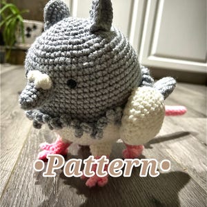 May include: Crocheted bird-like plush toy in gray, white, and pink. The handmade creature has a round body, small ears, and a beak-like feature. The word "Pattern" is displayed on the surface.