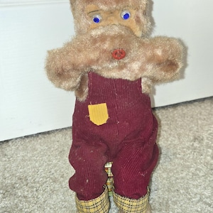 May include: A vintage plush toy with a fuzzy brown head, blue eyes, and a large brown mustache. It wears burgundy corduroy overalls with a yellow pocket and red shoes. The toy's legs have plaid cuffs.