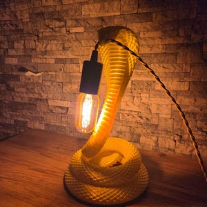 Pode incluir: Uma lâmpada amarela em forma de cobra com uma base enrolada e uma cabeça erguida. A lâmpada apresenta uma lâmpada de estilo vintage e um cabo preto. A lâmpada está colocada sobre uma superfície de madeira contra uma parede de tijolo.