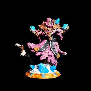 Könnte beinhalten: Eine bemalte Miniaturfigur eines Zauberers mit einem rosa Gewand und blauen Kristallen. Der Zauberer hält in jeder Hand einen blauen Kristall und steht auf einem Sockel mit blauen Kristallen.