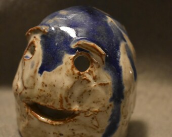 May include: A ceramic sculpture of a face with blue and brown glaze. The face has a hole for an eye and a mouth.