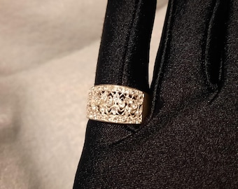 May include: A rectangular silver ring with a filigree design and small, clear gemstones. The ring is set against a black background, highlighting its intricate details and sparkle. A piece of jewelry.