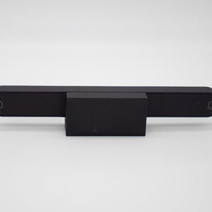 May include: A matte black horizontal bar component featuring a rectangular central block and two circular fasteners on the ends. The object has a clean, industrial design with a uniform dark finish and a symmetrical, elongated rectangular shape.