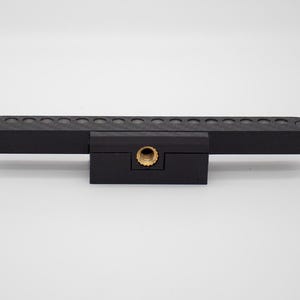 May include: A matte black horizontal mounting bar featuring a series of circular indentations along the top edge, secured by two screws at the ends, and a central brass-colored threaded insert for attachment purposes on the front face.