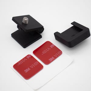 May include: A black plastic camera mount with a silver threaded screw, a separate black plastic mounting bracket, and two square red adhesive pads with the text 3M VHB printed in white, all arranged on a plain white surface.