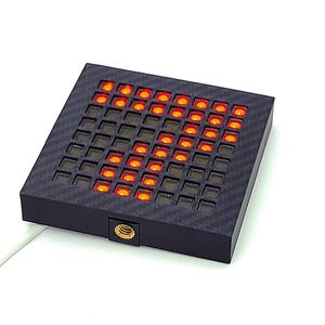 May include: A square, black electronic display with a grid of small, orange LED lights forming the number 7. The display has a textured surface and a small gold-colored screw on the side. A white cable extends from the device.
