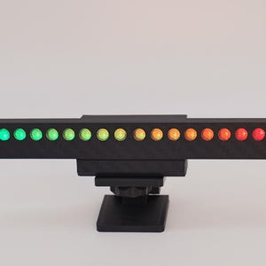 May include: A black horizontal bar device featuring a row of sixteen circular LED lights that transition in color from green on the left to yellow in the center and red on the right. The unit is mounted on a black base and has a white power cable.