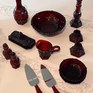May include: A collection of ruby red pressed glass tableware featuring a decanter with a stopper, a large bowl, a small bowl, a creamer, a butter dish with lid, two salt and pepper shakers, three candle holders, and two metal serving utensils.