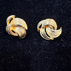 May include: A pair of gold-toned clip-on earrings. Each earring features a textured, swirling design with multiple curved elements. The earrings are set against a dark background, highlighting their metallic sheen.