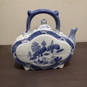 May include: A ceramic teapot featuring a blue and white color scheme with a hand-painted design of lotus flowers and two swimming ducks. The teapot has a curved handle, a small lid, and a spout, all decorated with blue floral patterns.