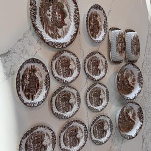 May include: A collection of vintage ceramic plates and serving dishes. The plates feature a brown and white transferware design with a landscape scene. The edges have a decorative, scalloped border. The set includes various sizes and shapes, mounted on a wall.