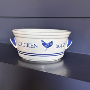 May include: A white ceramic soup bowl with two side handles, featuring blue text that reads CHICKEN SOUP and a blue graphic of a chicken in the center. The bowl is decorated with two horizontal blue stripes near the base and blue accents on the handles.
