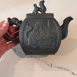 May include: Dark gray teapot with intricate relief carvings of a figure riding a tiger, a dragon-shaped handle, and a small statue on the lid. The teapot is made of a dark gray material and has a curved spout.